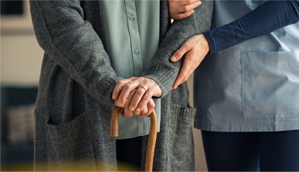 Vårdpersonal stödjer en äldre person med käpp.