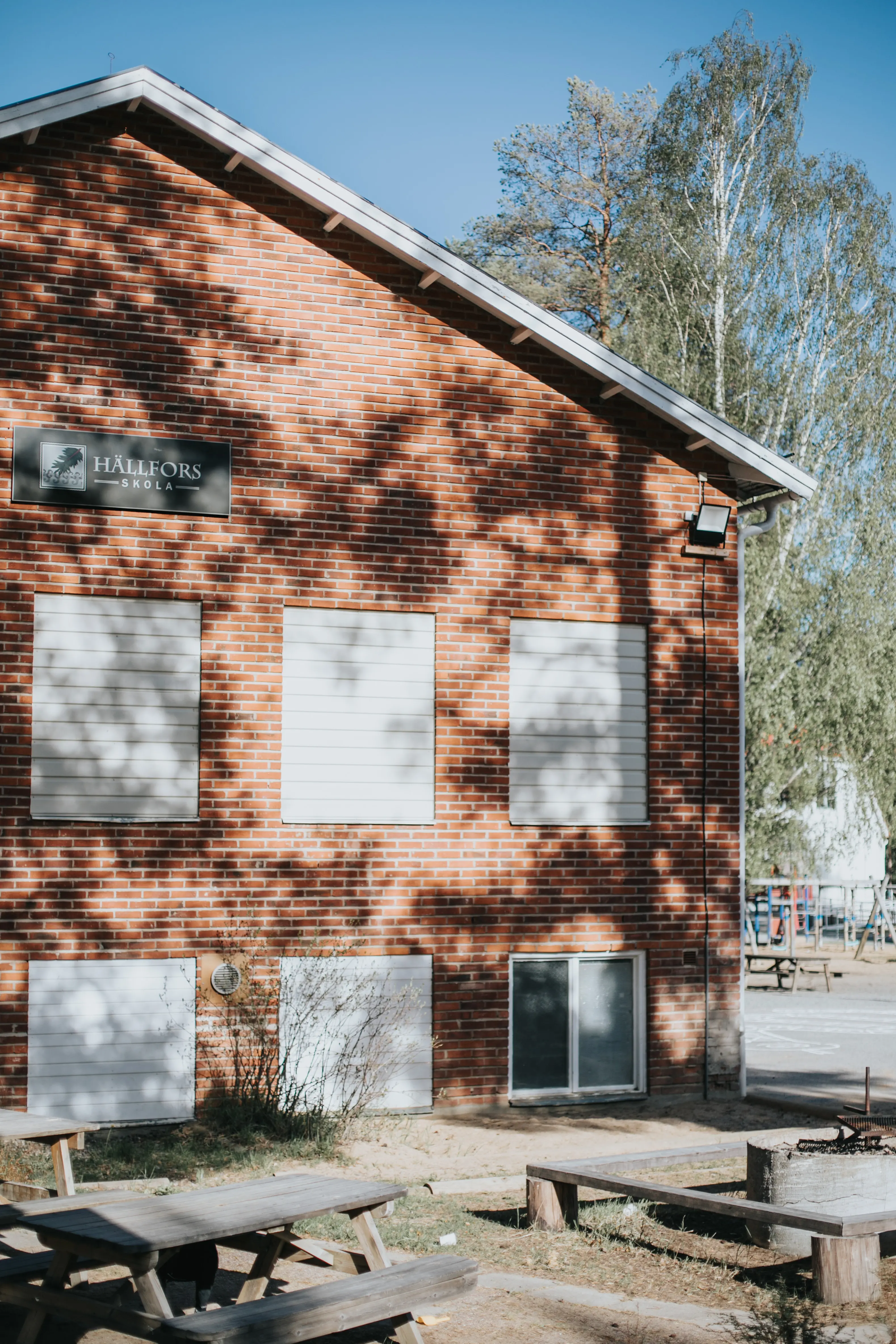 Gaveln på huvudbyggnaden i tegel vid Hällfors Friskola, grill-ring och bord med bänkar i förgrunden. Tallarna bakom fotografen skuggar lätt fasaden och silar solljuset.