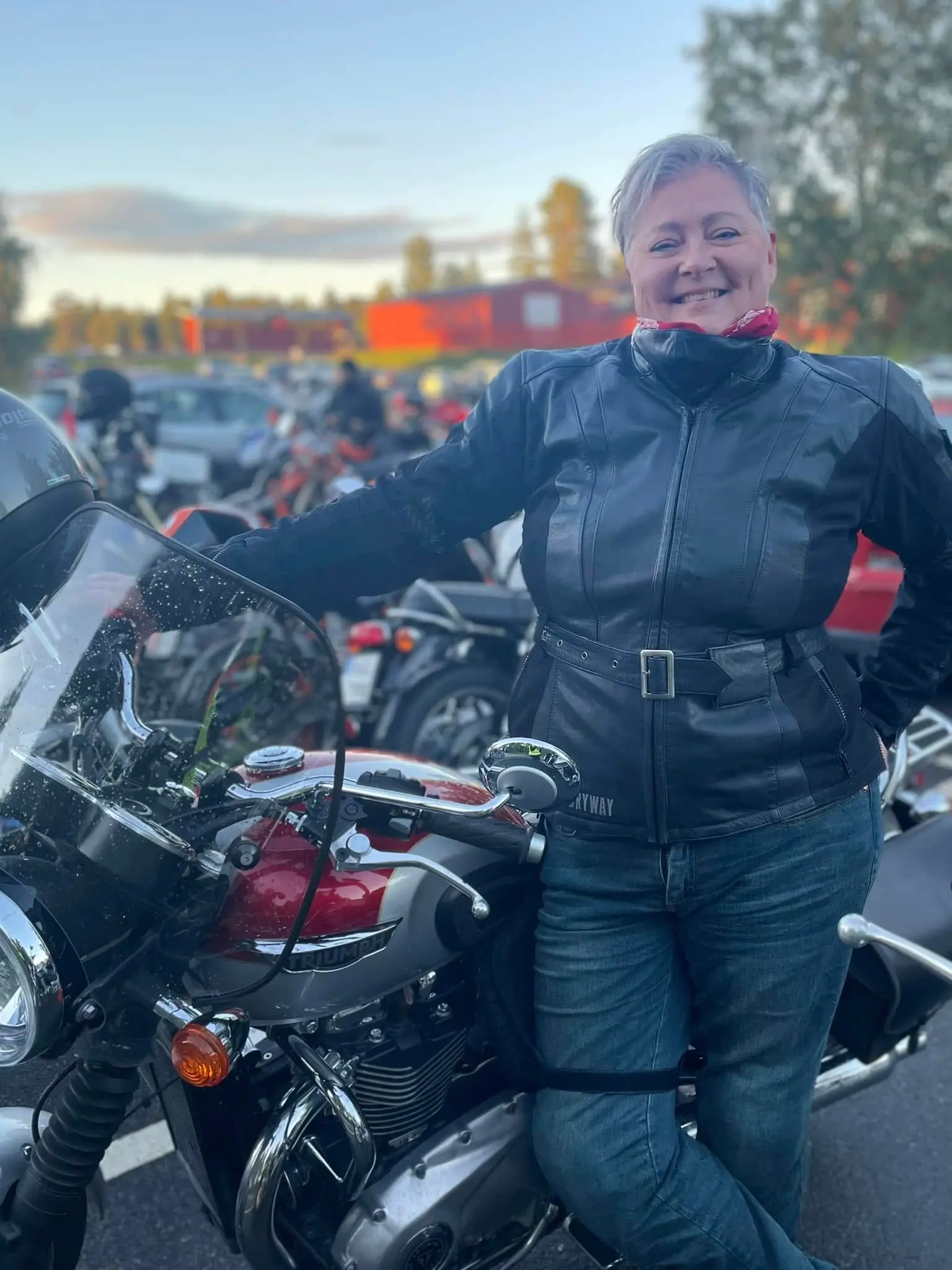 Patricia Riselius med ett stort brett leende, står i sitt MC-ställ vid sin Triumph MC på en MC-träff.