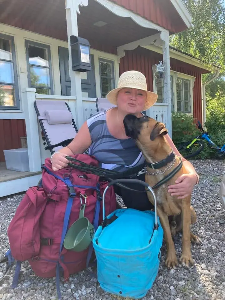 Patricia Riselius i stråhatt går ned på huk vid en vandringsryggsäck och kylväska när en av hennes hundar sitter intill och vill ge henne en puss.