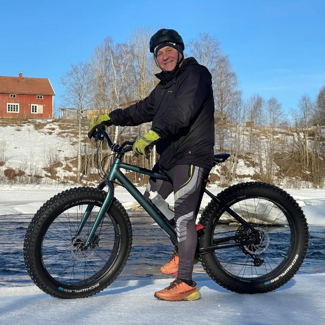 Anders Wonkavaara sportigt klädd, står över en Fatbike på en iskant med Vindelälven i bakgrunden.