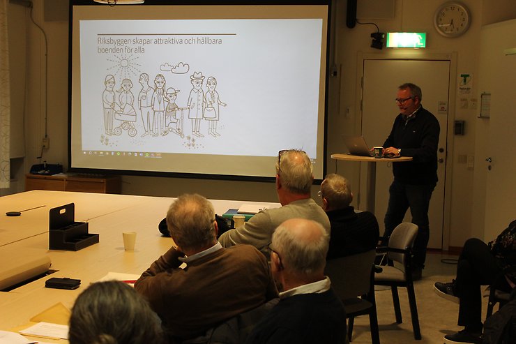 En man vid en projektorduk pratar inför personer som sitter vid bord.