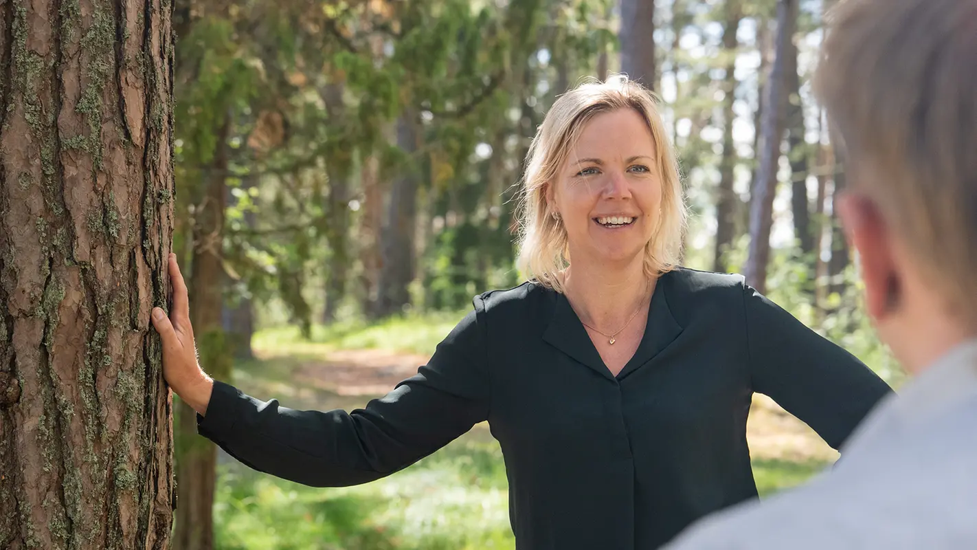 Glad kvinna i grön tröja lutar sig mot träd.
