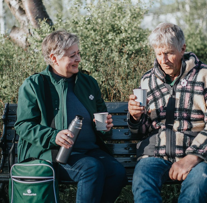 En kvinna med termos och en man på andra sidan av en parkbänk, båda med en kaffemugg i handen.